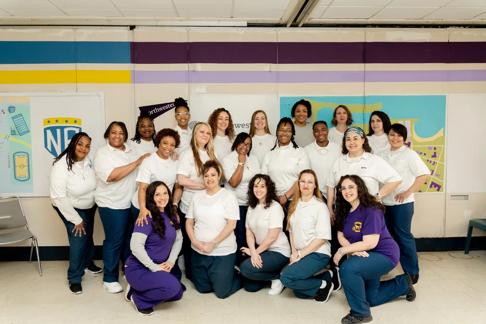 Group photo of about two dozen people, most in white shirts and dark pants, posed together in a classroom in front of a Northwestern pennant and a campus map mural.