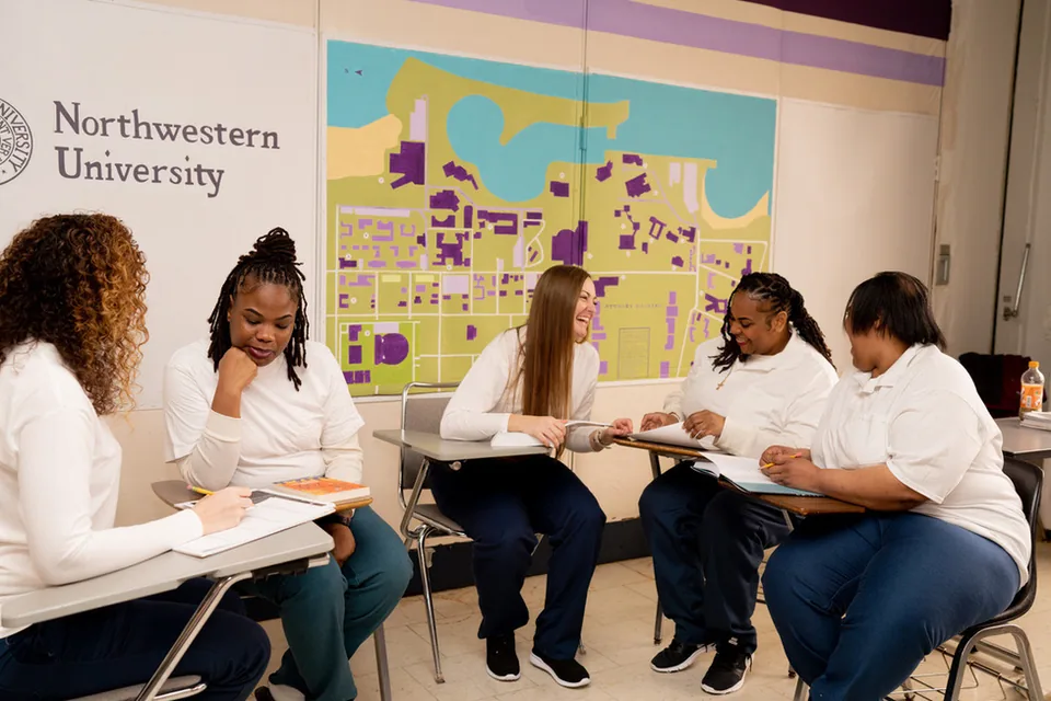 Three students in white shirts sit at classroom desks with open books in front of a campus map mural.