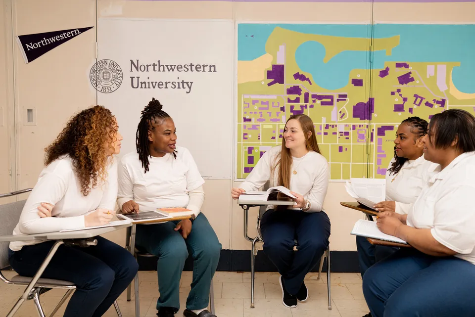 Two students sit at classroom desks with open books, one smiling toward the camera, beneath a wall reading “University” and a campus map mural.
