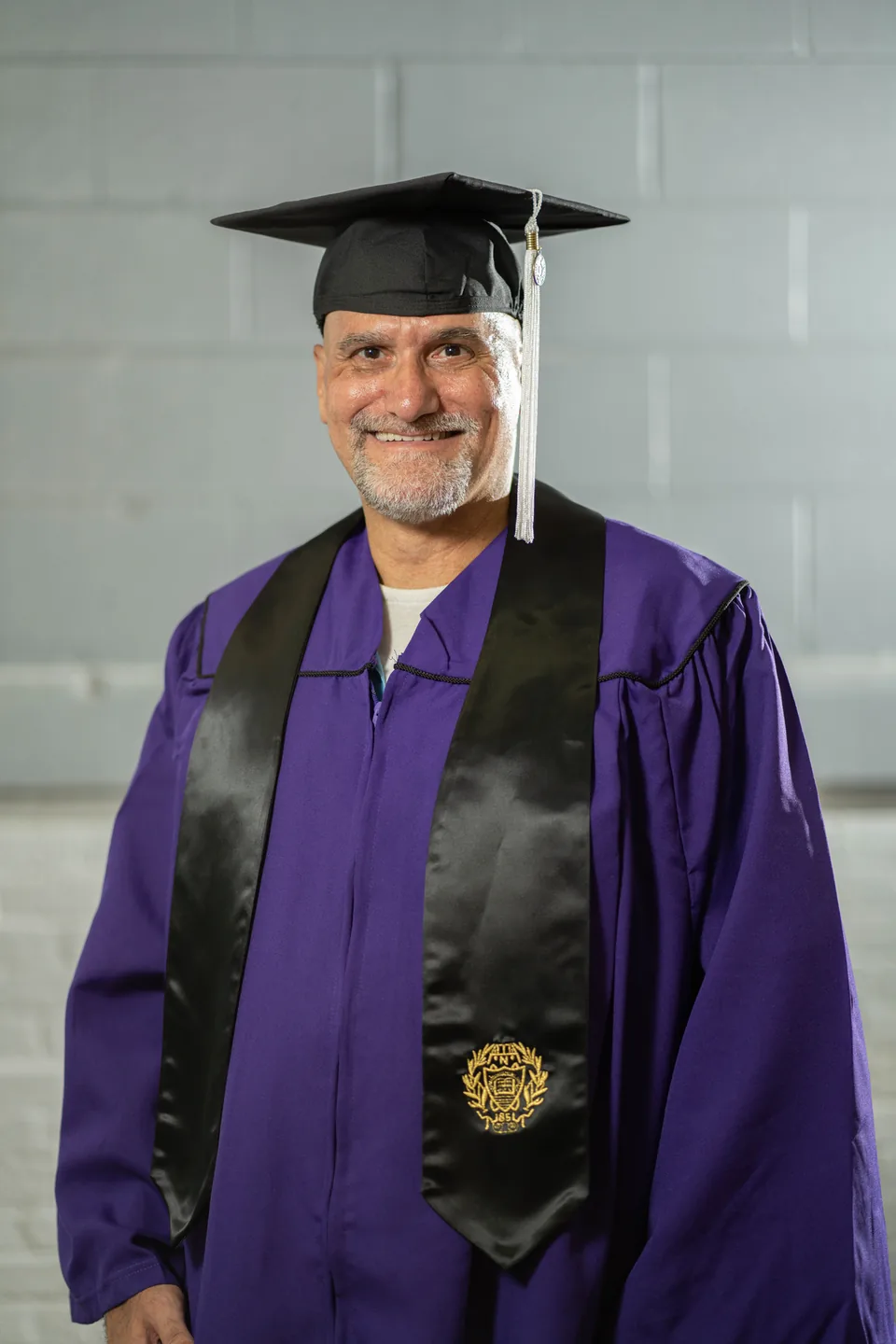Portrait of a smiling man with a gray goatee wearing a black graduation cap with silver tassel, a purple gown and a black stole.