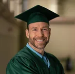 Headshot of a smiling man with a gray beard in a dark graduation cap and green gown.