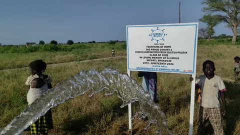 Water arcs from a pipe in the foreground of a grassy field where children stand beside a white sign reading "Fountains of Hope" in loving memory of alumnus Michael Broadway, sponsored by Embrace Relief.