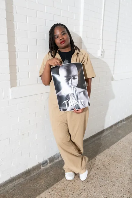 A woman in a tan prison uniform stands against a white brick wall, holding a black-and-white poster of Tupac Shakur.