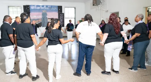 A group of people in black T-shirts and white or blue pants hold hands in a circle in a gymnasium-style room, with a stage backdrop and screens behind them.
