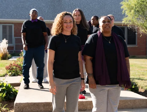 Several women in black T-shirts stand outdoors on a paved walkway in front of a brick building; two in front smile at the camera, one wearing a purple scarf.