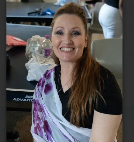 A smiling woman with long reddish-brown hair wears a black T-shirt and a white scarf with purple flowers, seated at a table.