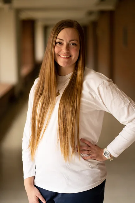 Photo of a smiling woman with long hair, wearing a white long-sleeved shirt and dark pants, standing with one hand on her hip in a hallway.