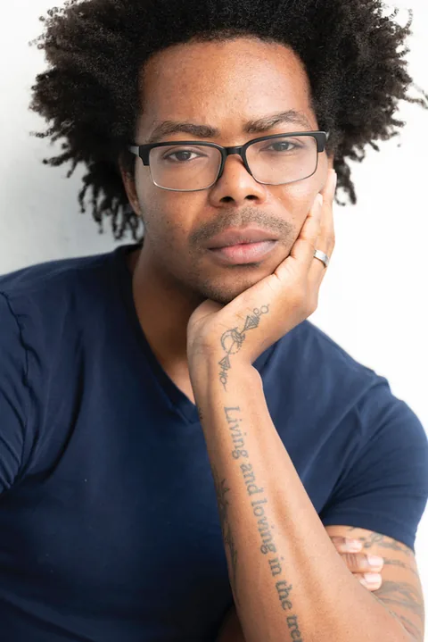 Close-up portrait of a person with curly hair and dark-rimmed glasses resting their chin on one hand; a forearm tattoo reads “Living and loving in the tre…” (cropped).