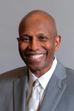 Head-and-shoulders portrait of Michael Deas, a smiling bald man in a gray suit jacket and open-collared light shirt, against a gray background.