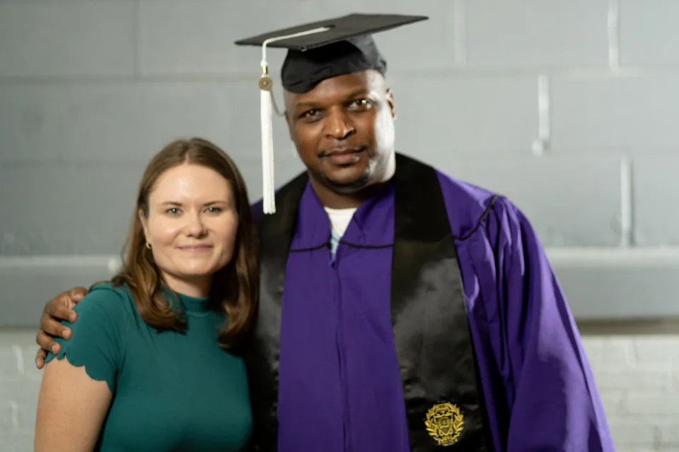 A smiling woman with long hair in a green top stands beside a man in a purple graduation gown and black mortarboard, his arm around her.