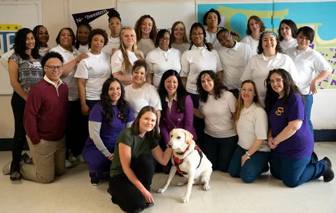 Group photo of about two dozen women, most in white shirts, posed in a classroom; a woman kneels in front with a yellow Labrador wearing a red harness.