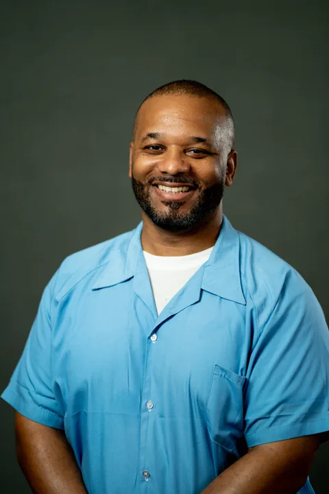 Studio portrait of Rommell Winters, a member of Cohort 5, in a light blue button-up shirt against a dark gray backdrop.