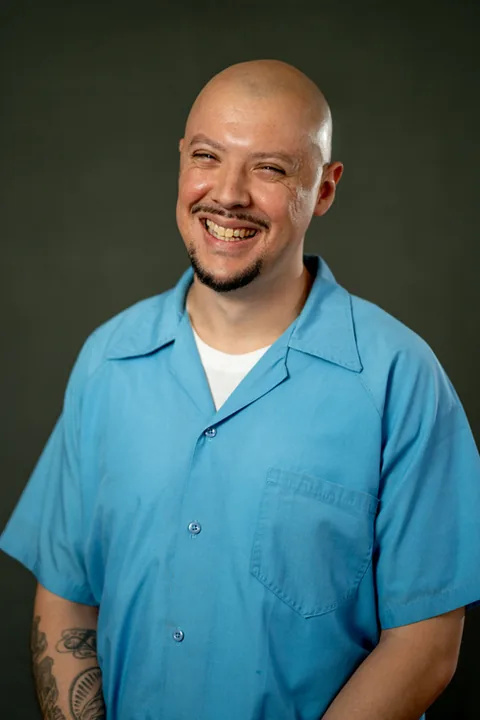 Studio portrait of Ariel Bueno, a member of Cohort 5, in a light blue button-up shirt against a dark gray backdrop.