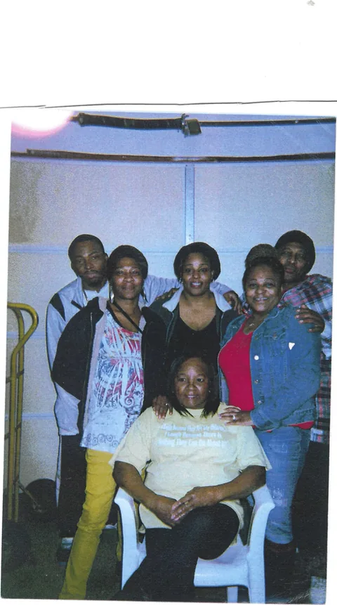 Snapshot-style family photo of seven adults posed in front of a garage door, one woman seated in a white plastic chair in front.