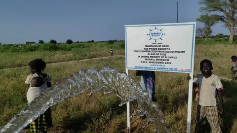 A jet of water arcs from the well pump in a grassy field while children stand nearby, next to a sign for the Fountains of Hope well built by NPEP Cohort 2 in memory of Michael Broadway.