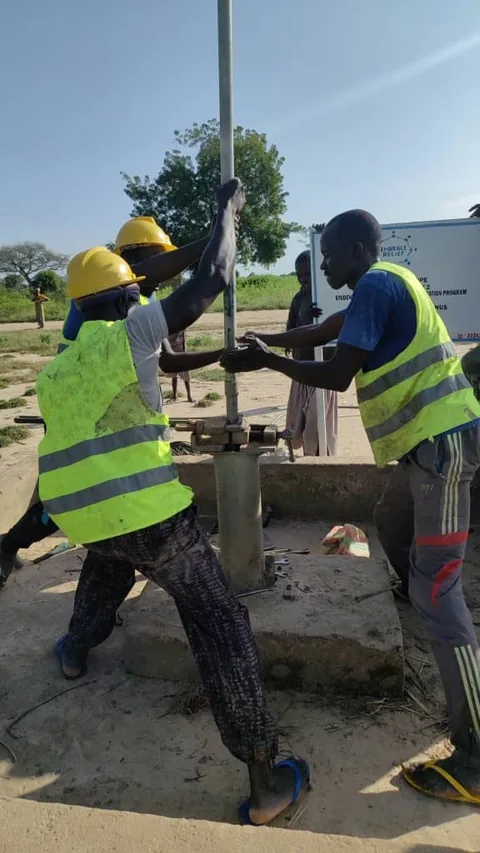 Workers in yellow hard hats and high-visibility vests grip a metal pipe as they install a well pump on a concrete base, with a sign behind them.