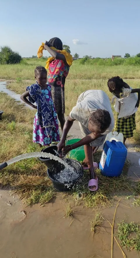 Girls collect water from a running hose into buckets and plastic jugs on muddy ground in a green field.