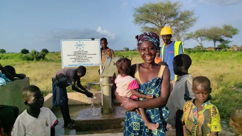 A smiling woman in a patterned dress holds a toddler beside the well while children and a worker in a hard hat stand around her and a boy washes at the pump.