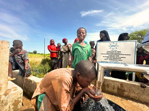 A boy bends to drink from his cupped hands under the well spout while children and women gather behind him near the Fountains of Hope sign.