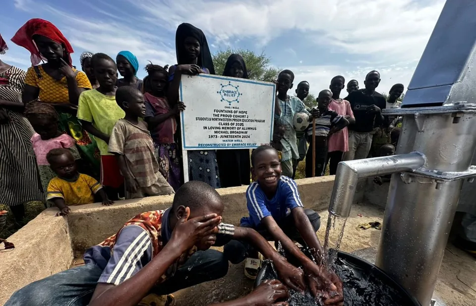 Two-page photo: children and villagers in Mailao, Chad, crowd around a new well and splash in its flowing water beside a sign reading "Fountains of Hope," dedicated in loving memory of Michael Broadway.