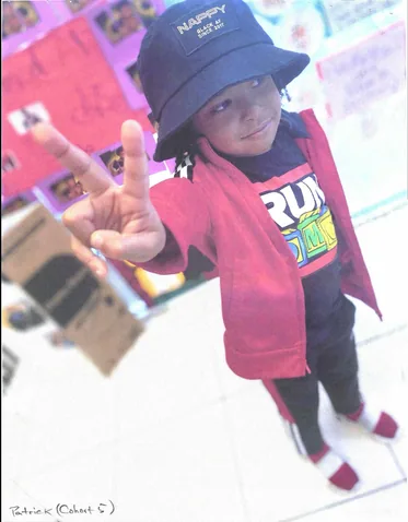 Photo of a young child in a bucket hat and red jacket making a peace sign.