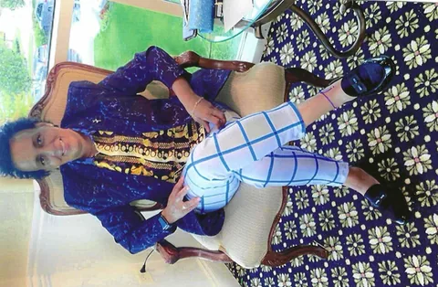 Photo of a woman in a sparkly blue jacket and patterned blouse sitting in an upholstered chair, legs crossed, in a decorated room.