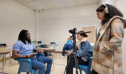 In a classroom, a man in a light blue shirt sits holding papers while a woman in a denim jacket sits beside a camera on a tripod; another person stands at right.