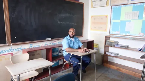 A man in a light blue shirt sits at a student desk in a classroom in front of a chalkboard, looking toward a window.