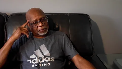 An older man with glasses and a gray T-shirt sits on a dark leather couch, resting his head on one hand as he speaks.
