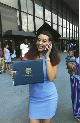 Photo of a smiling young woman in a graduation cap and blue dress holding a diploma cover and talking on a phone, outdoors.