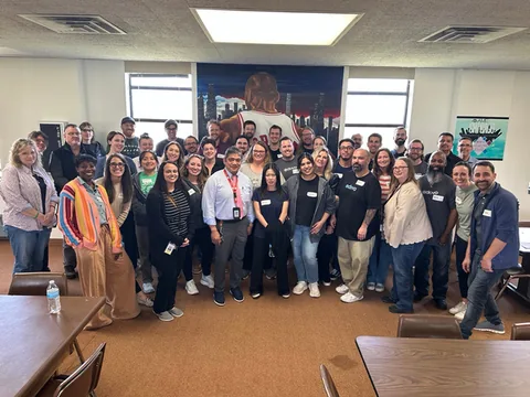 Large group photo of several dozen people posing together in a classroom-style room with tables and chairs.