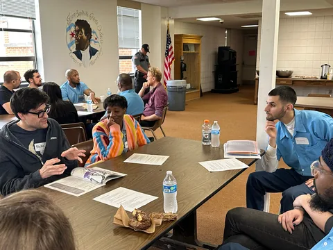 People seated around classroom tables in discussion, with water bottles and papers on the tables and a portrait and American flag on the wall behind them.