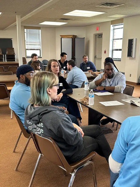 People seated around tables in a classroom, talking, with water bottles and papers in front of them.