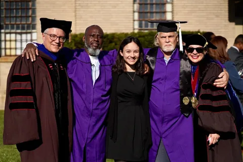 Five people stand together outdoors in front of a brick building; three wear academic robes and caps, two in purple gowns, and a young woman in a black dress stands in the middle.