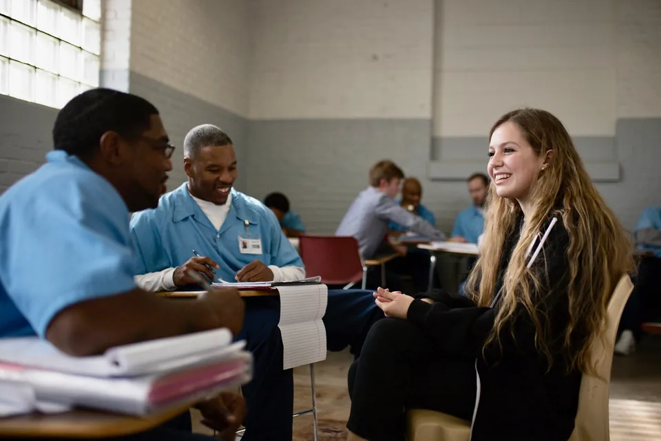 A classroom scene: a smiling young woman with long hair sits at a table across from men in light blue shirts, with papers and notebooks on the table and other people working in the background.