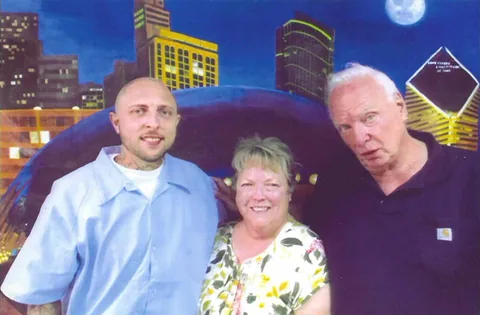 Photo of three people posing in front of a painted backdrop of a city skyline at night: a man in a light blue shirt, a smiling woman with short blond hair, and an older man with white hair in a dark shirt.